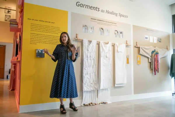 A woman stands in front of an exhibition titled "Garments as Healing Spaces," gesturing as she speaks. Behind her, white sensory garments for emotional regulation are displayed on a wall alongside descriptive text panels and photos illustrating their use.