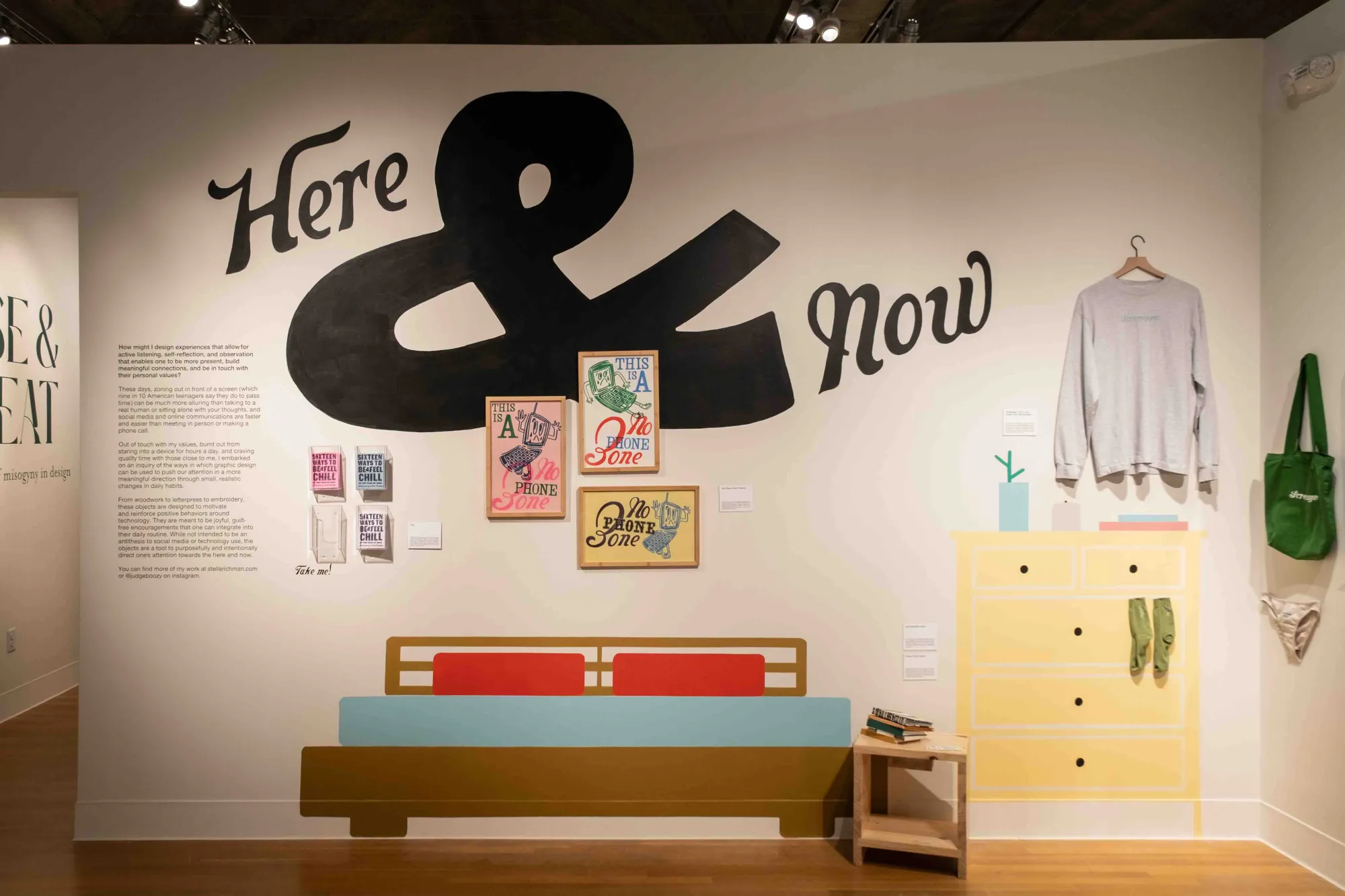 Installation view of a playful, home-themed design exhibit titled Here & Now. Wall art includes bold hand-painted text, “This is a No Phone Zone” prints, and whimsical objects like a sweatshirt, tote, and socks displayed against painted furniture. A statement about designing off-screen experiences is featured to the left.