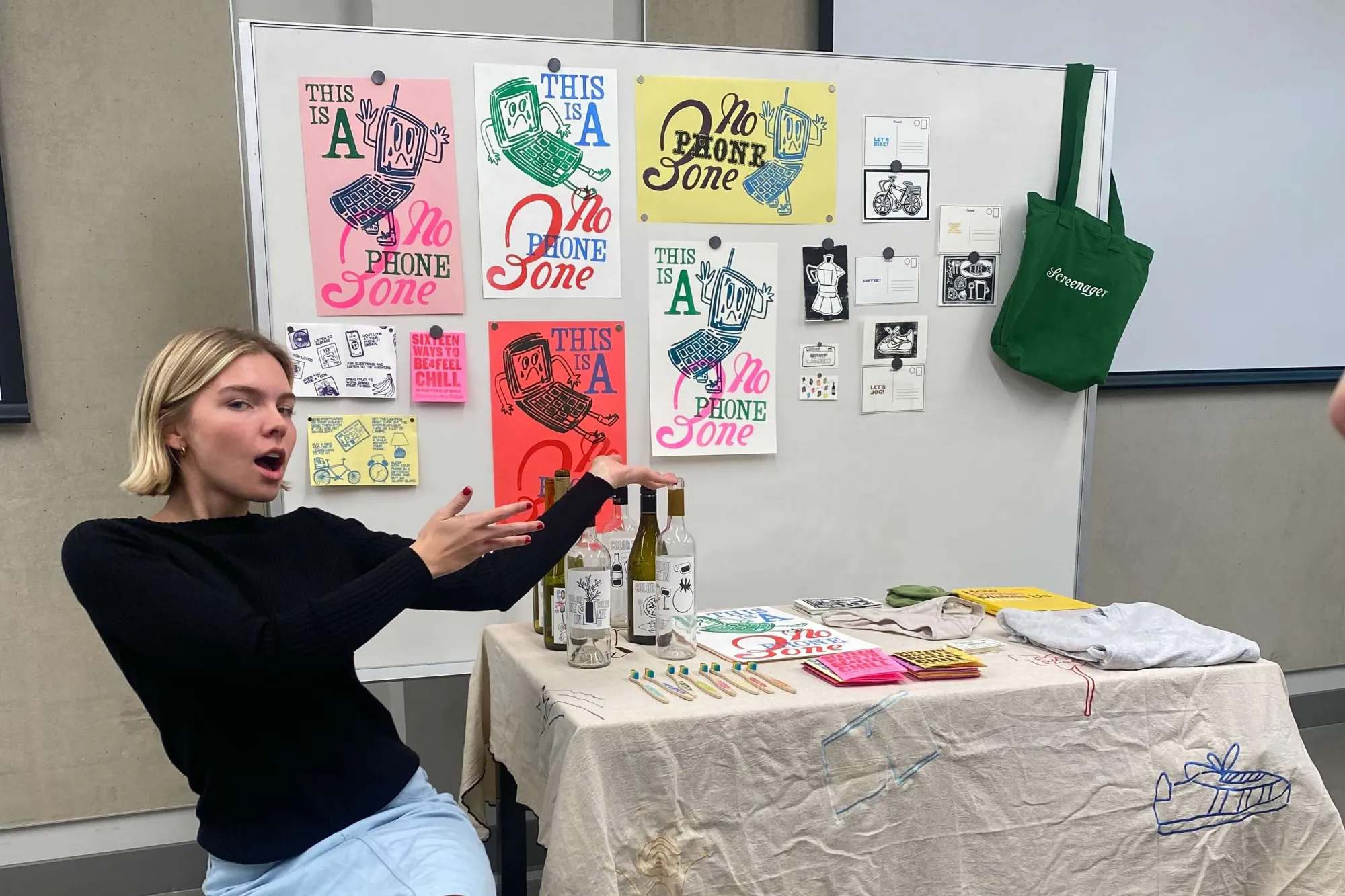 A student animatedly presents her thesis installation in a classroom setting, surrounded by vibrant risograph prints reading “This Is a No Phone Zone,” embroidered objects, illustrated bottles, and hand-labeled toothbrushes. A green tote bag and zine materials reinforce the theme of disconnecting from screens to reconnect with daily rituals.