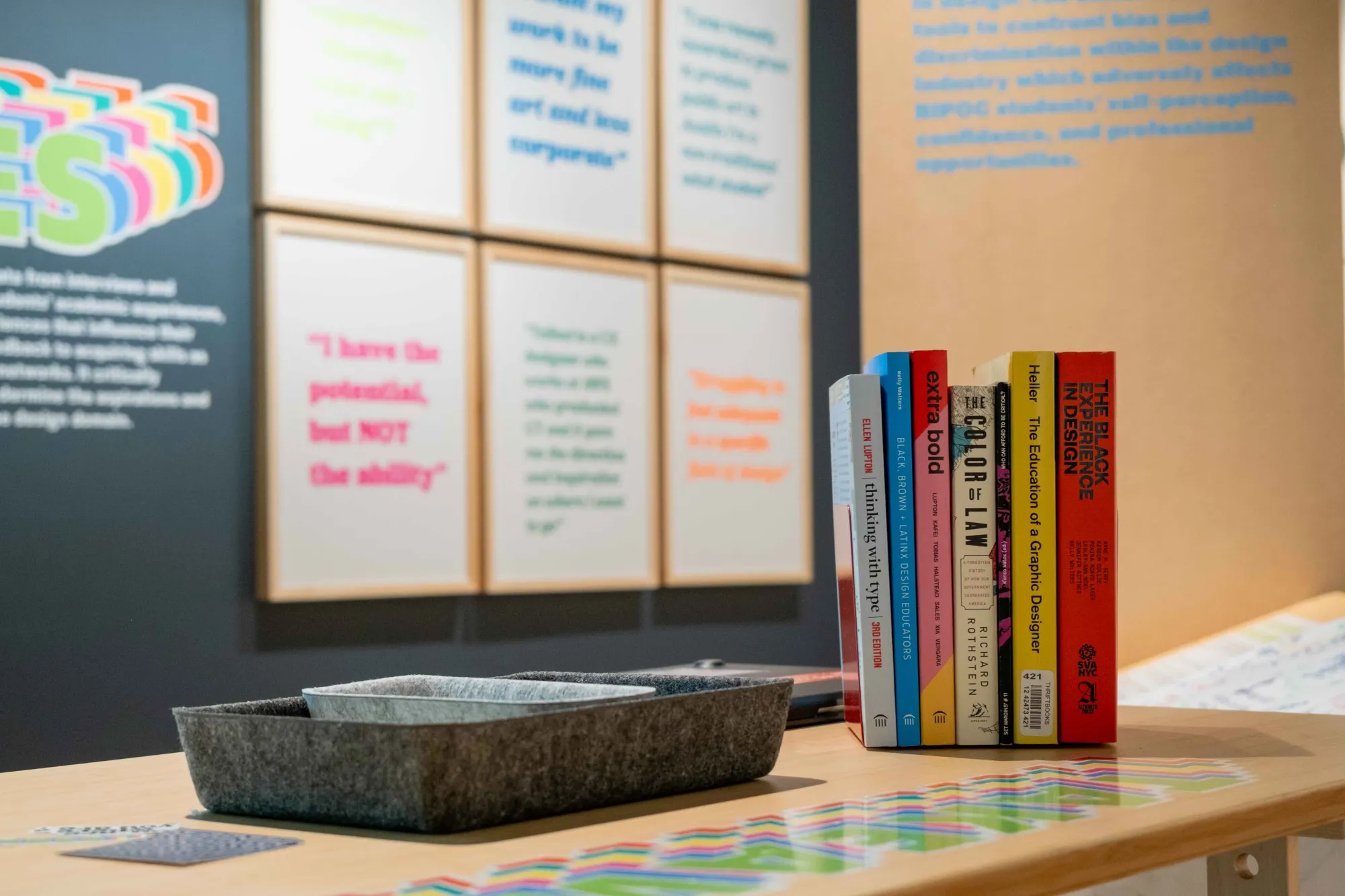 A gallery table displaying a collection of design books and printed materials, with framed student quotes and exhibition text panels visible on the wall behind.