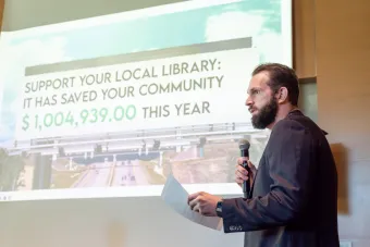 A bearded man in a navy blazer holds a microphone and paper while speaking in front of a projected slide. The slide displays a large billboard image with the text: “SUPPORT YOUR LOCAL LIBRARY: IT HAS SAVED YOUR COMMUNITY $1,004,939.00 THIS YEAR.” The background shows a highway overpass and a partly cloudy sky.