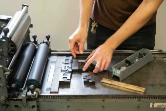Close-up of a person arranging large wooden letterpress type blocks on an old-fashioned printing press. Their hands are positioning the letters while a tray of additional type rests nearby.