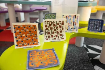 Colorful embroidered textile swatches with graphic patterns are displayed upright on a lime green table at a design exhibition. The background shows additional multicolored tables and exhibit materials.