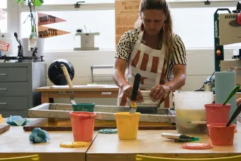 An instructor wearing a striped apron prepares materials at a printmaking station, surrounded by colorful ink containers, spatulas, and printing tools. A “Wood Type” pennant hangs in the window behind her, emphasizing a hands-on studio environment.