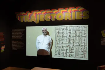 A person stands smiling in front of a projected display featuring a maze artwork, beneath a large colorful typographic installation reading "Trajectories" at a design exhibition.