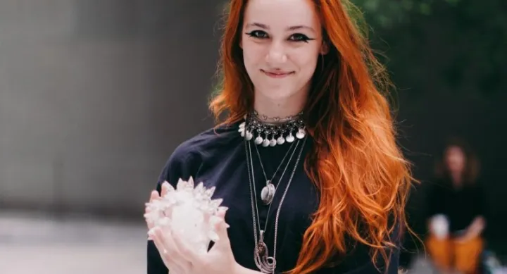 Person with long red hair and layered silver jewelry holds a translucent crystal sculpture outdoors, smiling warmly while looking directly at the camera. The background is softly blurred with greenery and another person in the distance.