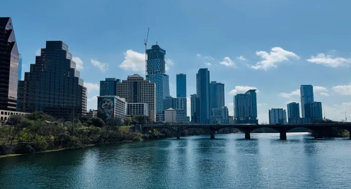 A wide view of the Austin skyline with high-rise buildings, the Colorado River in the foreground, and a bridge crossing the water.