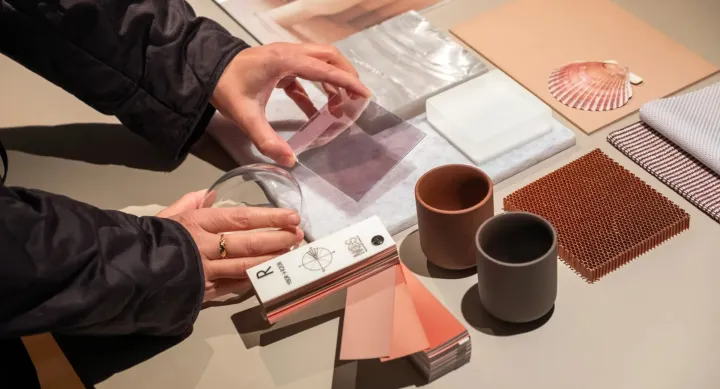 A person’s hands interact with a variety of material samples arranged neatly on a table. The samples include translucent acrylic sheets, color swatches, ceramic cups, fabric swatches, a shell, and textured tiles in neutral and earthy tones. The person holds a clear circular lens and a square plastic sheet, suggesting a material exploration or design planning activity.