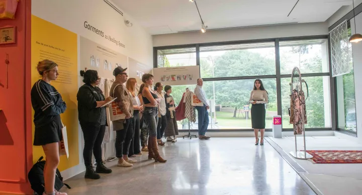 A group of people stand in a gallery space listening to a woman speak near large windows. The exhibition, titled "Garments as Healing Spaces," features text panels, sensory garments, and clothing installations focused on emotional regulation and care.