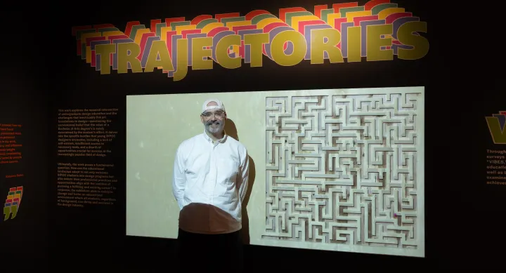 A person stands smiling in front of a projected display featuring a maze artwork, beneath a large colorful typographic installation reading "Trajectories" at a design exhibition.